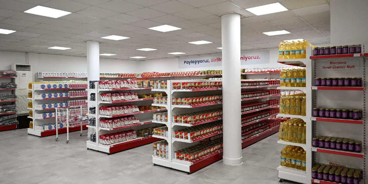 Modern bir market iç mekanında, raflarda düzenli bir şekilde dizilmiş gıda ürünleri yer alıyor. Raflarda sıvı yağ şişeleri, konserve kutuları ve çeşitli paketli gıda ürünleri bulunuyor. En sağdaki raf ünitesinde 