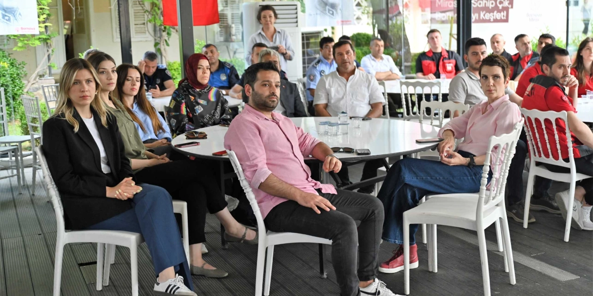 Açık havada, beyaz yuvarlak masaların etrafında oturmuş, dikkatle dinleyen bir grup insan. Ön planda siyah ceketli bir kadın, pembe gömlekli bir erkek ve açık pembe gömlekli başka bir kadın yer alıyor. Arka planda ise farklı kıyafetler giymiş, toplantıyı takip eden katılımcılar ve bazı görevliler bulunuyor. Ortam, bir eğitim veya bilgilendirme toplantısını andırıyor.