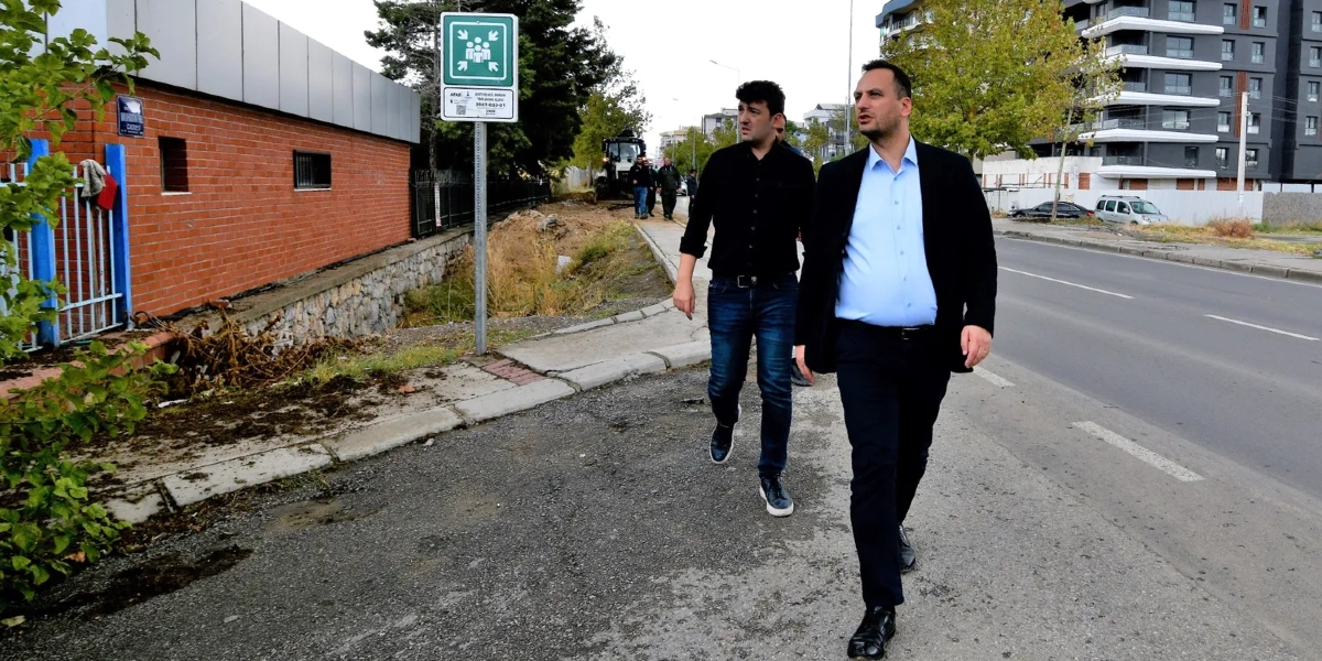 Bornova'da bir yol kenarında yürüyen, biri siyah ceketli diğeri siyah gömlekli iki erkek. Yanlarında AFAD acil toplanma alanı tabelası bulunuyor ve arka planda inşaat çalışmaları ile modern binalar görülüyor.