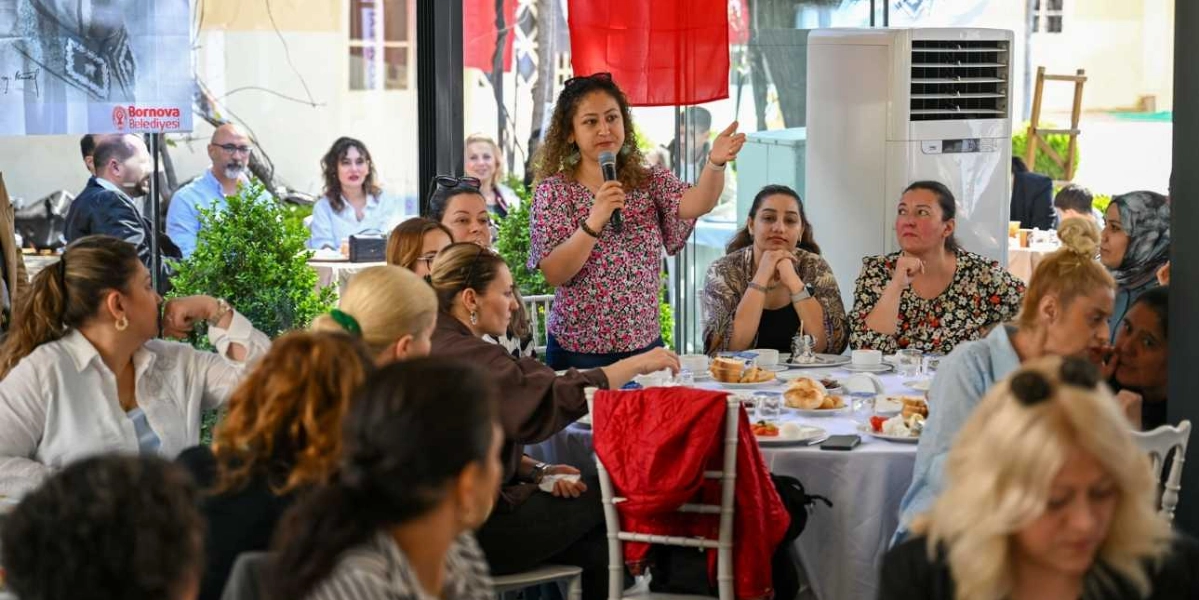 Bornova Belediyesi tarafından düzenlenen bir etkinlikte, çiçek desenli bluz giyen bir kadın ayakta mikrofonla konuşma yaparken, masalarda oturan katılımcılar onu dinliyor. Kapalı bir alanda gerçekleşen toplantıda, beyaz örtülü masaların etrafında toplanan kadınlar ve erkekler yer alıyor. Arka planda Bornova Belediyesi logosunun bulunduğu bir afiş ve ayakta duran bir klima cihazı görülüyor.