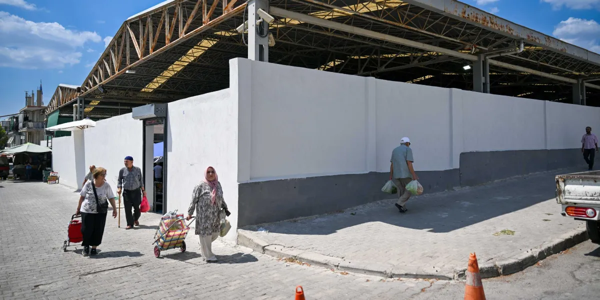 Üstü kapalı, beyaz duvarlarla çevrili bir pazaryerinin dış cephesinde, ellerinde pazar poşetleri ve tekerlekli pazar arabalarıyla yürüyen insanlar görülüyor. Pazaryerinin giriş kapısının üzerinde güvenlik kamerası yer alıyor. Gün ışığında çekilmiş karede, kaldırımda yürüyen vatandaşlar ve sağ tarafta park halindeki bir kamyonetin arka kısmı dikkat çekiyor.