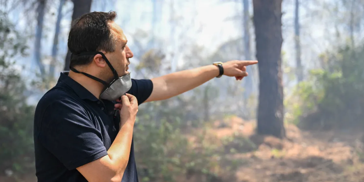 Koyu renkli polo yaka tişört giymiş, yüzünde koruyucu maske bulunan bir erkek, ormanlık alanda dumanların yükseldiği bir noktayı eliyle işaret ederek yönlendirme yapıyor.