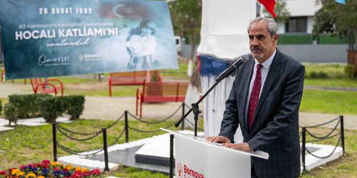 Bornova Belediyesi kürsüsünde konuşma yapan gri saçlı ve bıyıklı bir erkek, arka planda üzerinde 