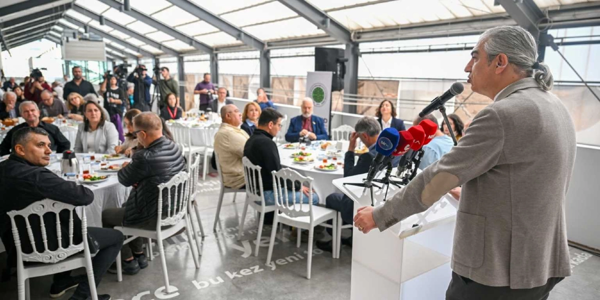 Modern, cam tavanlı ve aydınlık bir salonda, kürsüde konuşma yapan gri ceketli ve arkadan bağlı saçlı bir erkek. Kürsüde çeşitli haber kanallarının mikrofonları bulunuyor. Salonda yuvarlak masalarda oturan ve konuşmacıyı dinleyen bir grup davetli yer alıyor. Zemin üzerinde büyük harflerle yazılmış metinler dikkat çekiyor.