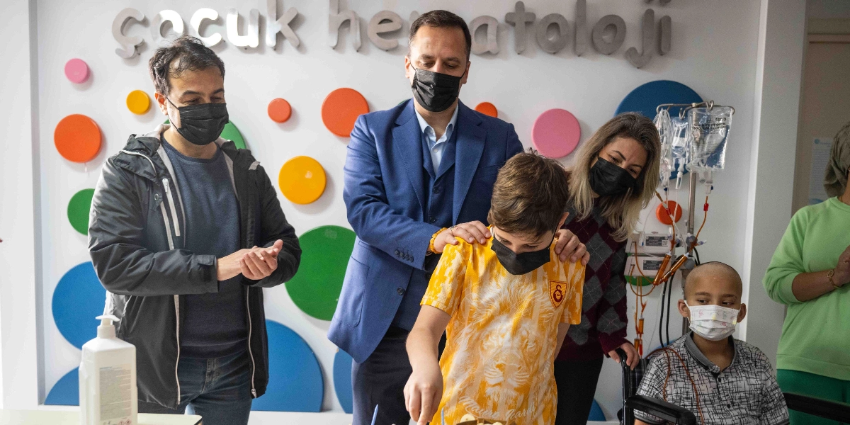 Çocuk Hematoloji servisinde, üzerinde renkli daireler bulunan bir duvarın önünde, maskeli yetişkinler ve çocuklar bir arada duruyor. Sarı Galatasaray forması giyen bir çocuk, önündeki pastayı keserken, arkasında duran mavi ceketli bir adam ellerini çocuğun omuzlarına koymuş destek veriyor. Yanlarında maskeli bir kadın ve diğer yetişkinler onları izlerken, sağ tarafta tekerlekli sandalyede oturan ve maske takan başka bir çocuk daha bulunuyor.