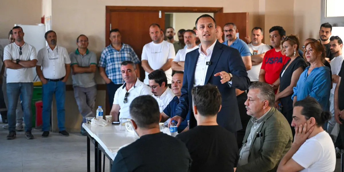Lacivert ceketli bir adam, kapalı bir mekanda masanın başında ayakta durarak konuşma yapıyor. Etrafında, çoğu erkeklerden oluşan kalabalık bir grup, dikkatle onu dinliyor. Bazı kişiler masada otururken, diğerleri ayakta duruyor. Ortam, resmi olmayan bir toplantı veya çalışma alanı havası taşıyor.