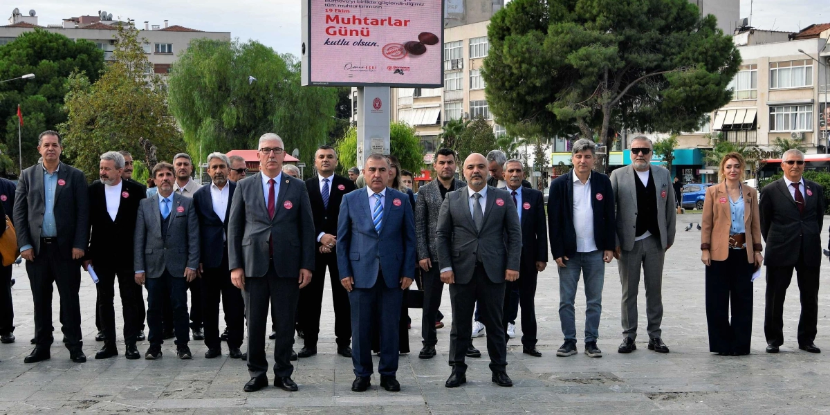 Bornova'da bir meydanda, 19 Ekim Muhtarlar Günü kutlaması kapsamında bir araya gelen, çoğunluğu takım elbiseli erkeklerden ve bir kadından oluşan grup, arkalarında üzerinde 