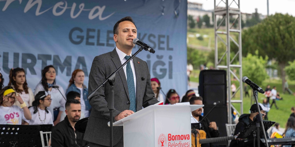 Koyu gri takım elbiseli ve desenli kravatlı bir erkek, üzerinde Bornova Belediyesi logosu bulunan bir kürsüde mikrofon başında konuşma yapıyor. Arka planda 