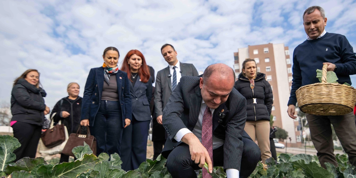 CHP Genel Başkan Yardımcısı Özgür Karabat, Bornova'da bir bahçede çömelmiş vaziyette sebze hasadı yaparken, çevresinde yerel yöneticiler ve vatandaşlar onu izliyor. Karabat'ın sağ tarafında elinde hasat sepeti tutan bir erkek, sol tarafında ise kadın ve erkeklerden oluşan bir grup bulunuyor. Arka planda ise çok katlı bir apartman ve bulutlu bir gökyüzü yer alıyor.