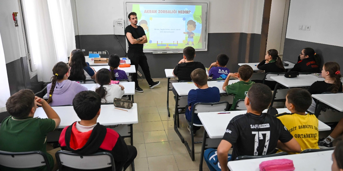 Sınıf ortamında, siyah tişörtlü bir eğitmen tahtanın önünde durarak öğrencilere akran zorbalığı hakkında sunum yapıyor. Akıllı tahtada 