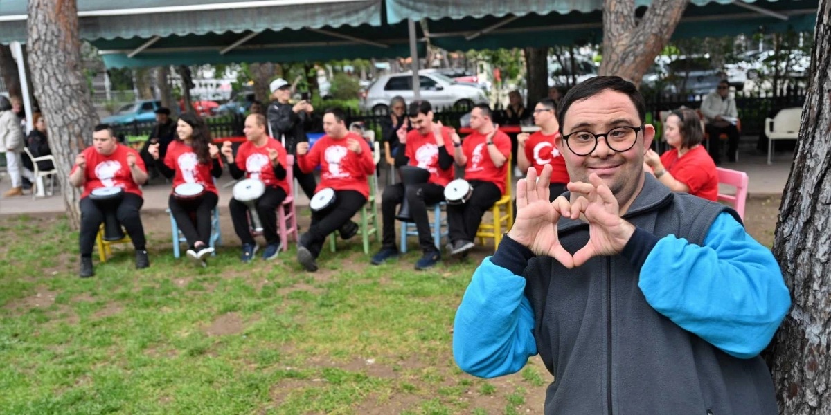 Açık havada, ağaçların altında oturan ve ellerinde darbuka bulunan kırmızı tişörtlü bir grup Down sendromlu genç, ön planda elleriyle kalp işareti yapan gözlüklü bir gençle birlikte neşeli bir şekilde poz veriyor.