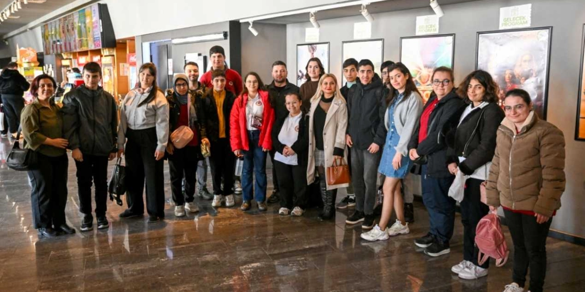 Bir sinema salonunun fuayesinde yan yana duran, farklı yaş gruplarından oluşan kalabalık bir öğrenci ve öğretmen grubu. Gruptaki kişiler günlük kıyafetler ve kışlık montlar giymiş, gülümseyerek kameraya bakıyorlar. Arka planda duvarda asılı film afişleri ve 