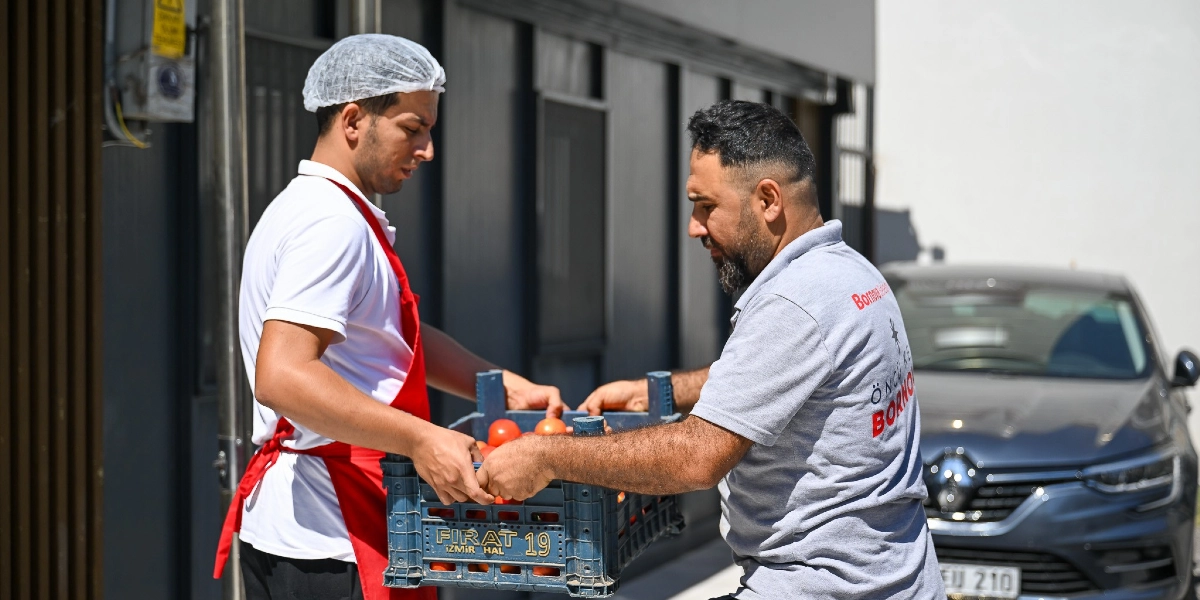 Beyaz önlüklü, başında bone takan bir çalışan ile üzerinde Bornova Belediyesi logolu gri bir tişört bulunan diğer bir görevli, içinde domatesler bulunan plastik bir kasayı birlikte taşıyor.