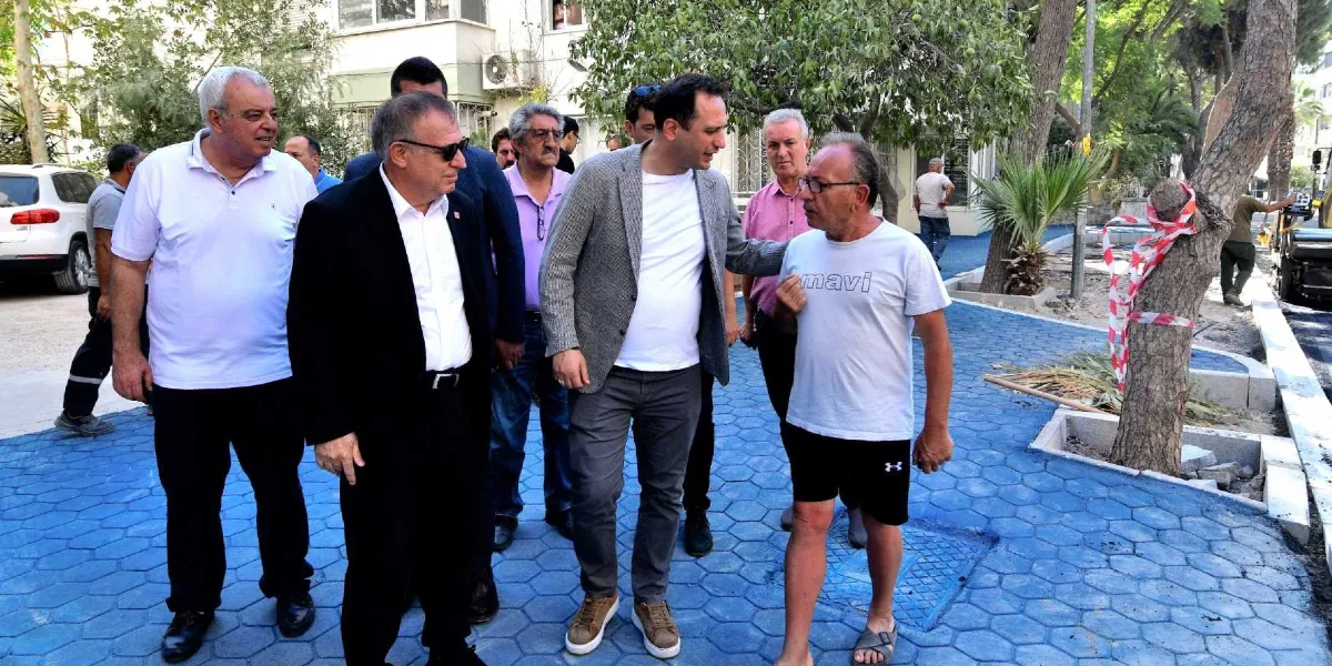 Yenilenmekte olan 159 Sokak'ta, mavi renkli altıgen taşlarla döşenmiş yeni kaldırım üzerinde yürüyen bir grup erkek, devam eden çalışmaları inceliyor. Grupta yer alan kişiler arasında beyaz tişörtlü, ceketli ve takım elbiseli yetkililer bulunuyor; bir kişi ise üzerinde 