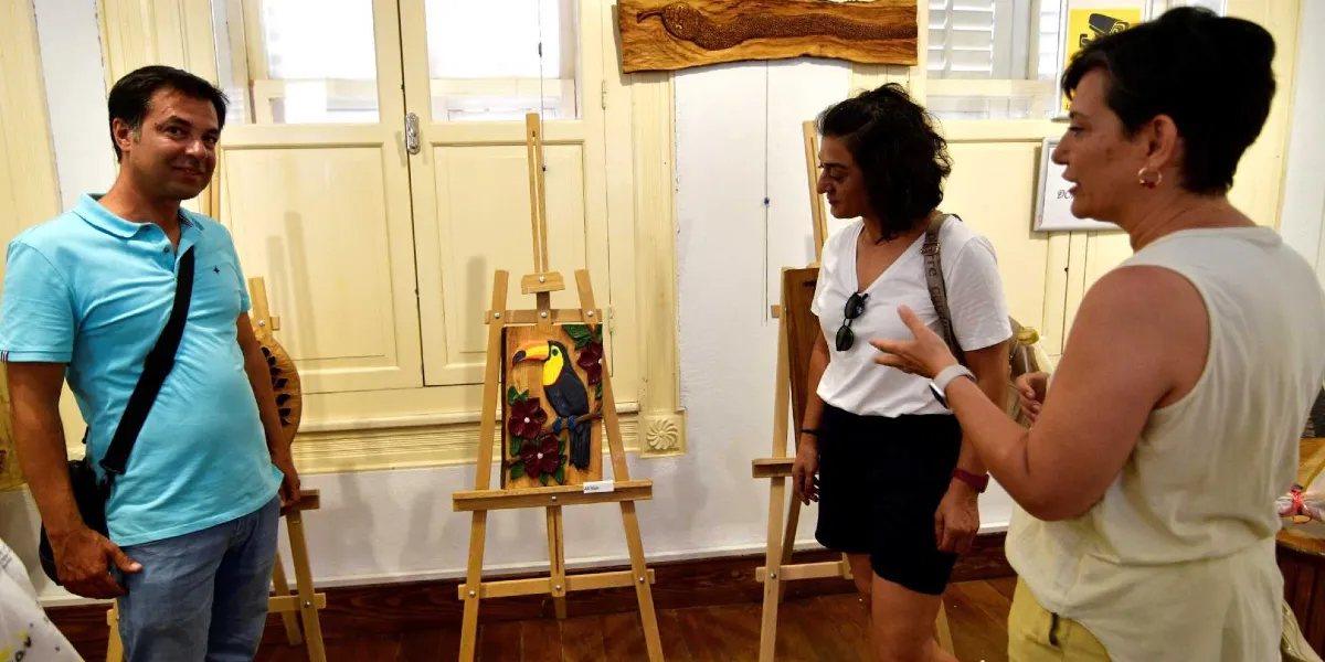 Aydınlık bir sanat galerisinde, ahşap şövaleler üzerinde sergilenen oyma ahşap eserleri inceleyen üç kişi. Sol tarafta açık mavi polo yaka tişörtlü bir erkek, ortada beyaz V yaka tişörtlü ve siyah şortlu bir kadın, sağ tarafta ise beyaz kolsuz bir bluz giyen başka bir kadın yer alıyor. Ortadaki şövalede, renkli detaylara sahip bir tukan kuşu figürünün işlendiği ahşap bir tablo bulunuyor. Arka planda ise beyaz ahşap pencereler ve duvarda asılı başka bir ahşap oyma eser görülüyor.