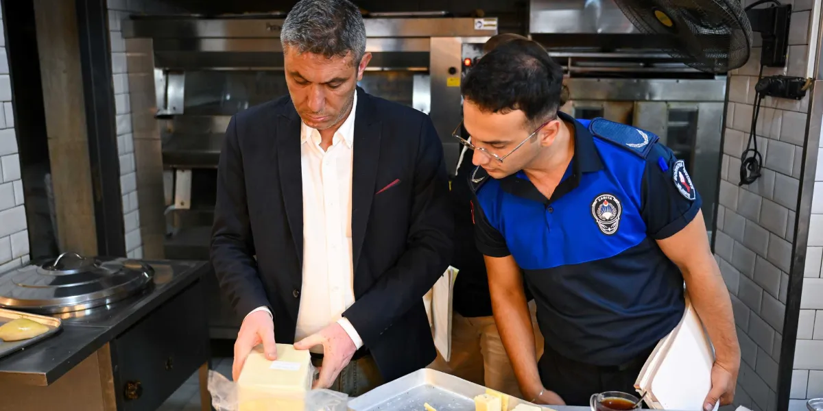 Koyu renk ceket ve beyaz gömlek giymiş bir yetkili ile Bornova Belediyesi zabıta üniformalı bir görevli, bir fırın mutfağında tezgah üzerindeki gıda ürünlerini inceliyor. Zabıta memurunun kolunda Bornova Belediyesi arması bulunuyor, arka planda ise endüstriyel fırınlar ve mutfak ekipmanları yer alıyor.