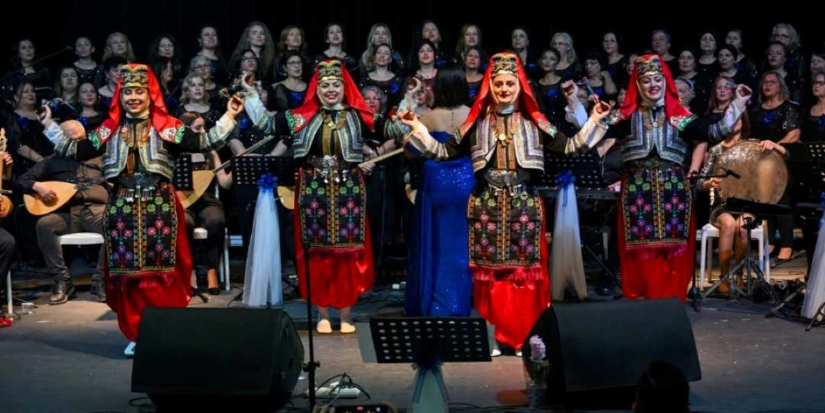 Sahne üzerinde geleneksel kıyafetler giymiş dört kadın halk dansçısı, ellerinde küçük zillerle dans ederken görülüyor. Kırmızı başörtülü ve işlemeli yelekli dansçıların arkasında, koyu renkli kıyafetler giymiş kalabalık bir kadın korosu ve enstrüman çalan müzisyenler yer alıyor. Sahnenin önünde iki büyük hoparlör ve nota sehpaları bulunuyor.