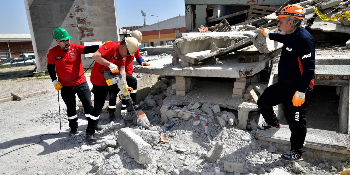 Enkaz alanında arama kurtarma eğitimi yapan ekip üyeleri; kırmızı tişörtlü ve baretli görevliler kırıcı delici aletle beton parçalarını kırmaya çalışırken, AFAD logolu kıyafet giyen ve başında kask bulunan bir görevli çalışmaları izliyor.