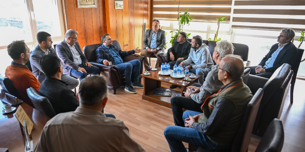 Ahşap panellerle kaplı bir ofis ortamında, deri koltuklarda oturan ve bir toplantı gerçekleştiren on kişilik erkek grubu. Ortadaki alçak sehpanın üzerinde su şişeleri ve kahve fincanları bulunuyor. Grup üyeleri birbirleriyle sohbet ederken, bazıları dikkatle konuşmacıyı dinliyor. Oda aydınlık olup, pencerelerin önünde yeşil bitkiler ve jaluzi perdeler yer alıyor.