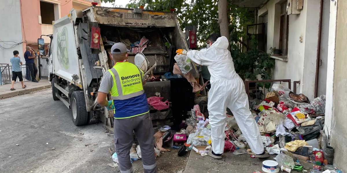 Bornova Belediyesi Fen İşleri Müdürlüğü yeleği giyen bir görevli ile beyaz koruyucu tulumlu bir başka görevli, bir binanın önünde birikmiş yoğun çöp yığınını çöp kamyonuna yüklüyor.