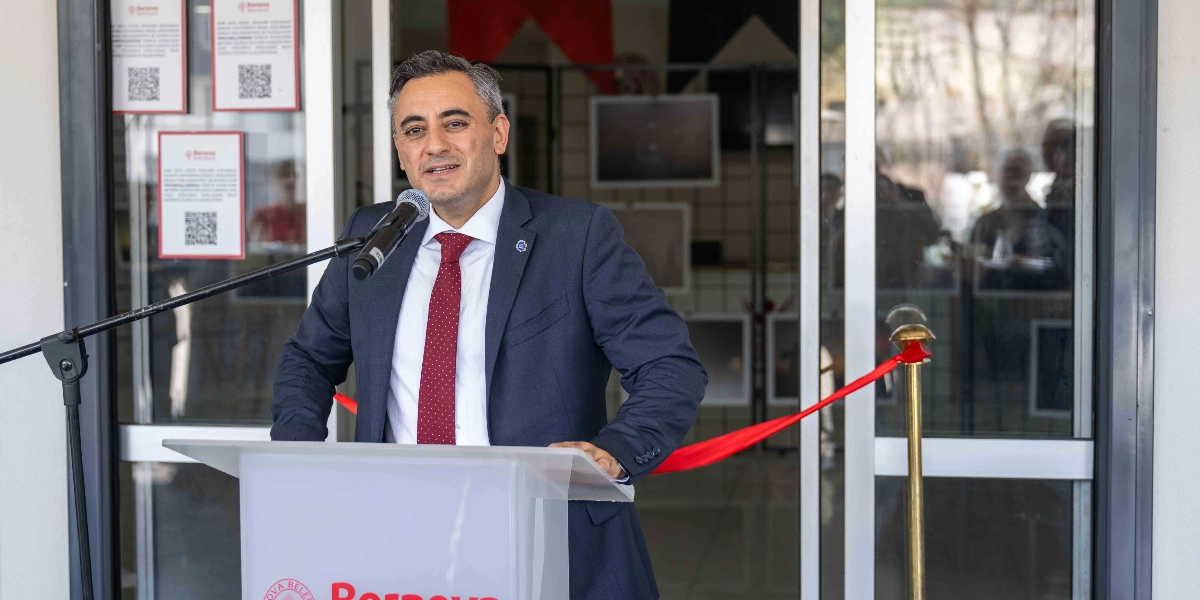 Lacivert takım elbiseli ve kırmızı kravatlı bir erkek, Bornova Belediyesi logosunun bulunduğu bir kürsüde mikrofon başında konuşma yapıyor. Arka planda cam kapılı bir giriş ve üzerinde Bornova Belediyesi logolu QR kod içeren bilgilendirme panoları yer alıyor. Kapının önünde açılış törenlerini simgeleyen kırmızı bir kurdele şeridi bulunuyor.
