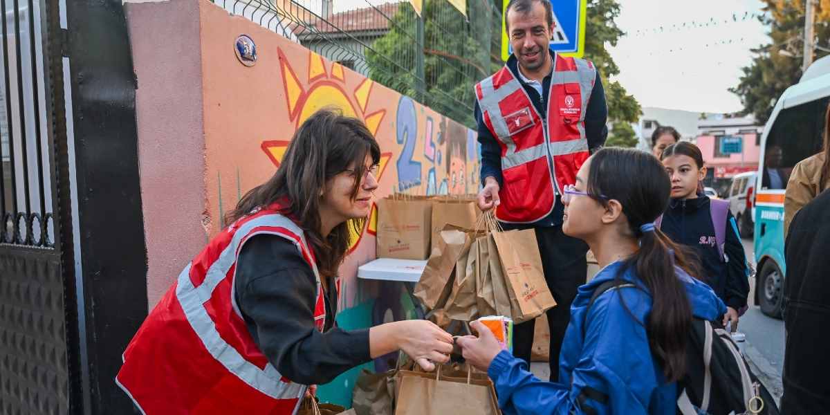Bornova Belediyesi logolu kırmızı yelekler giymiş iki görevli, okul çıkışında öğrencilere üzerinde Bornova Belediyesi yazan kahverengi kağıt beslenme çantalarını dağıtıyor. Görevlilerden biri bir öğrenciye çanta uzatırken, diğer görevli elindeki çantaları hazırlıyor. Arka planda duvarında güneş ve çocuk figürleri olan renkli bir okul duvarı ile okul servisi aracı görünüyor.