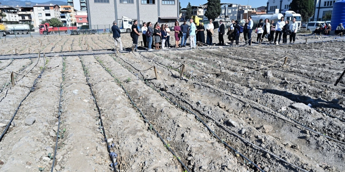 Açık havada, yeni sürülmüş ve damlama sulama boruları döşenmiş geniş bir tarım arazisinde bir grup insan yan yana durarak alanı inceliyor. Arka planda şehir binaları, park halindeki tırlar ve sanayi yapılarının bulunduğu bir yerleşim alanı görülüyor.