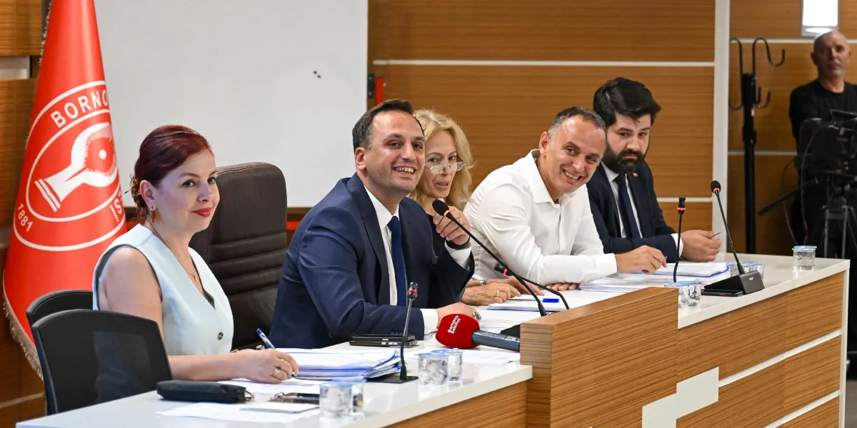 Bornova Belediyesi'nin düzenlediği bir toplantıda, uzun bir masada yan yana oturan beş kişilik bir heyet yer alıyor. Masanın en solunda Bornova Belediyesi'nin kırmızı renkli logosunun bulunduğu bir bayrak duruyor. Heyetteki kişiler, önlerinde bulunan mikrofonlar ve dosyalarla bir toplantı veya basın açıklaması gerçekleştiriyor. Katılımcılar arasında Bornova Belediye Başkanı Ömer Eşki de bulunuyor; kendisi lacivert takım elbiseli, gülümseyerek konuşuyor. Masanın diğer ucunda ise bir kamera operatörü çekim yapıyor.