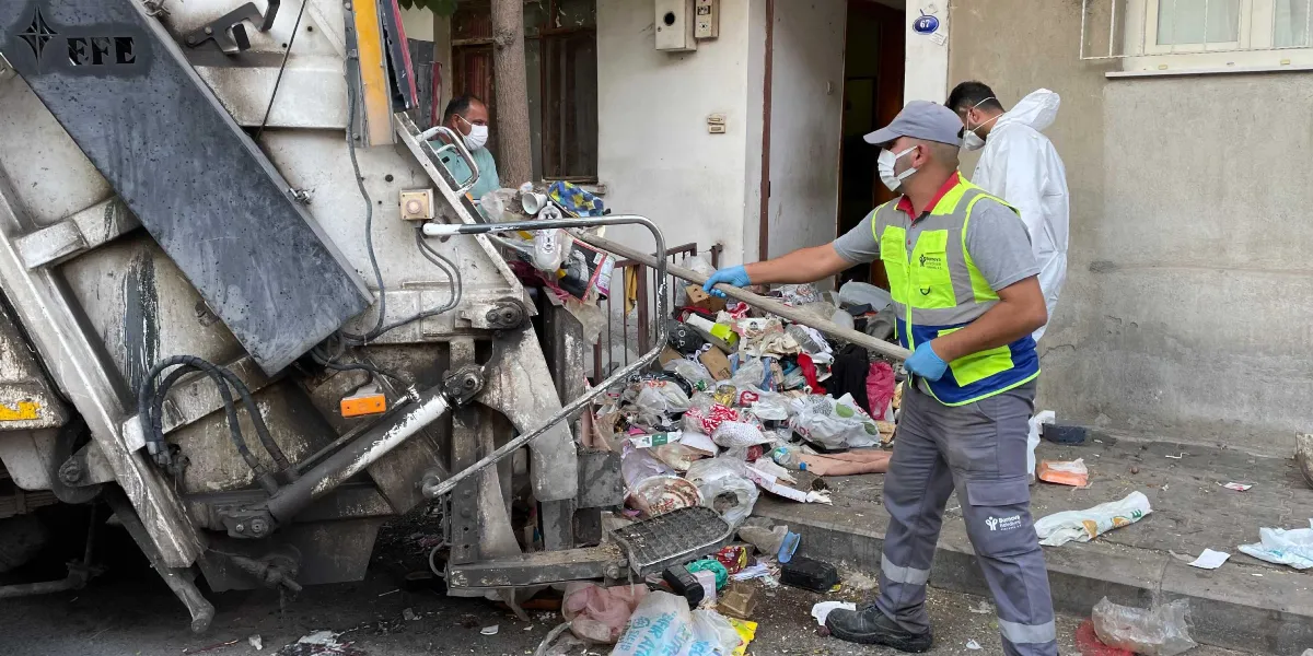 Bornova Belediyesi temizlik görevlileri, bir apartman girişinde birikmiş olan yoğun çöp yığınını temizliyor. Maskeli ve eldivenli görevliler, ellerindeki süpürge ve küreklerle çöpleri çöp kamyonuna yüklüyor; arka planda beyaz koruyucu tulum giymiş bir başka görevli daha bulunuyor.