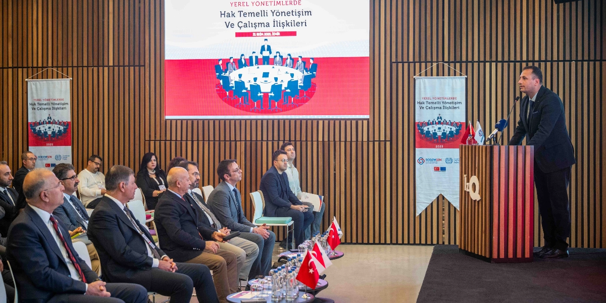 Modern bir konferans salonunda kürsüde konuşma yapan bir yetkili ve onu dinleyen katılımcılar yer alıyor. Arka plandaki büyük ekranda ve yanlardaki dikey afişlerde 