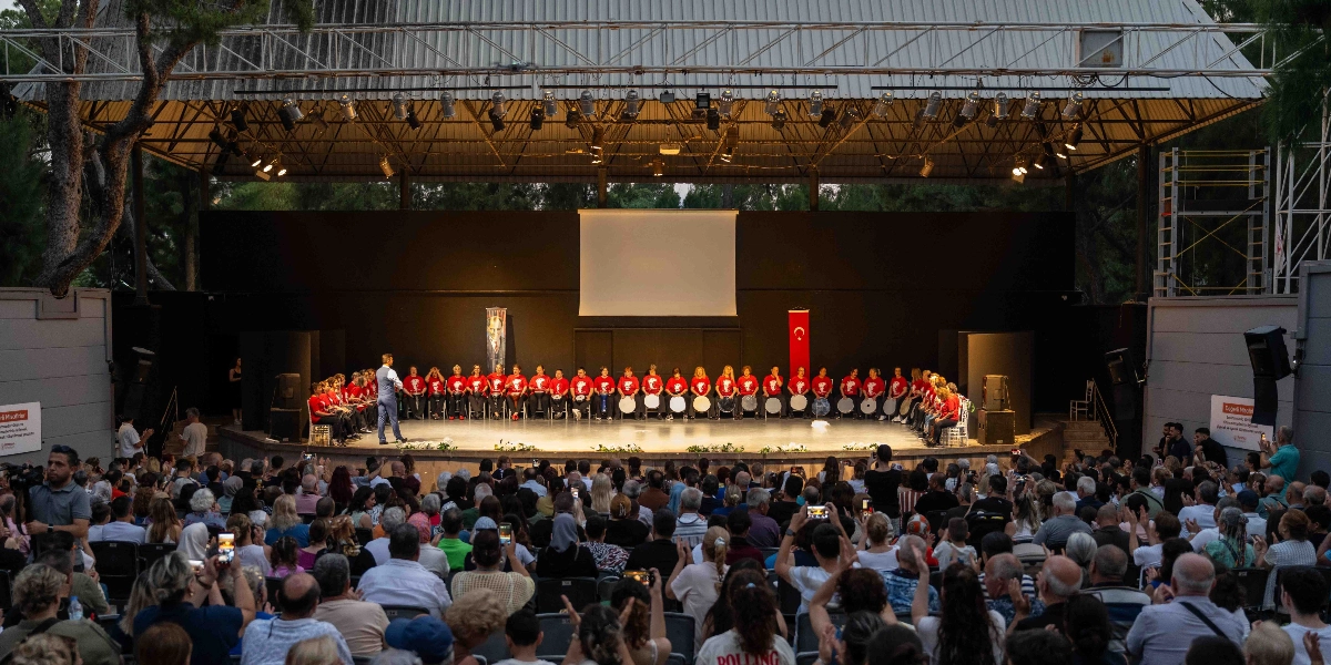 Açık hava sahnesinde, kırmızı tişörtlü ve ellerinde davul bulunan geniş bir grup müzisyen, bir şef yönetiminde performans sergiliyor. Sahnenin arka planında beyaz bir perde, Atatürk posteri ve Türk bayrağı yer alıyor. Sahne önünde, etkinliği izleyen ve alkışlayan kalabalık bir seyirci grubu bulunuyor.