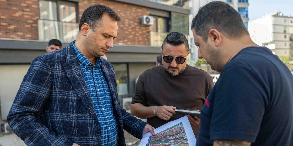 Üç erkek, dış mekanda bir arada durarak ellerindeki kentsel planlama projesini veya haritayı inceliyor. Mavi kareli gömlek ve lacivert ceket giyen kişi haritayı tutarken, yanındaki güneş gözlüklü kişi elindeki tabletten projeyi takip ediyor, üçüncü kişi ise onları izliyor. Arka planda modern bir apartman binası yer alıyor.
