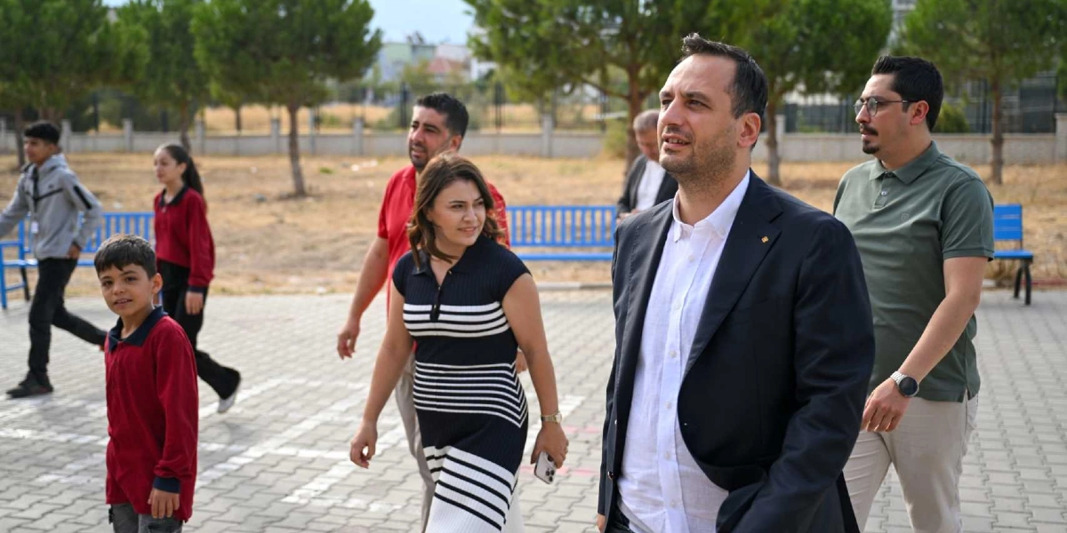 Açık havada, okul bahçesi olduğu anlaşılan parke taşlı bir alanda yürüyen bir grup yetişkin ve arka planda hareket halindeki öğrenciler. Ön planda koyu renkli bir ceket ve beyaz gömlek giymiş bir erkek, yanında siyah beyaz çizgili bir elbise giyen bir kadın ve haki renkli polo yaka tişörtlü bir erkek yer alıyor. Arka planda kırmızı tişörtlü bir erkek, okul forması giymiş öğrenciler ve mavi banklar bulunuyor.