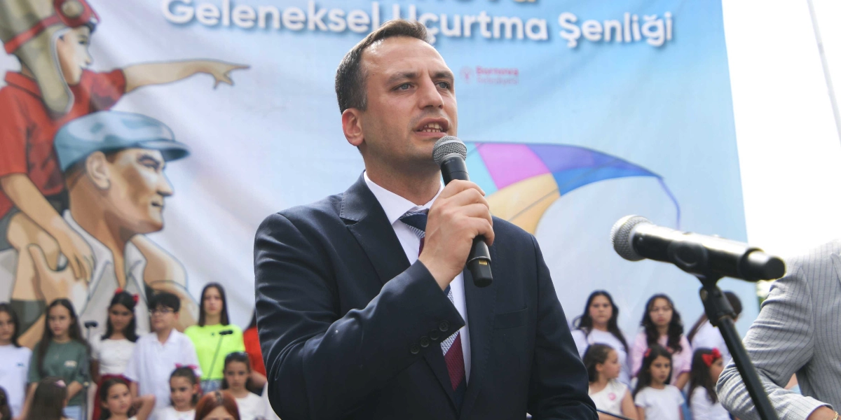 Koyu renk takım elbiseli bir erkek, elinde mikrofonla konuşma yapıyor. Arka planda Bornova Belediyesi'nin düzenlediği Geleneksel Uçurtma Şenliği yazılı bir pankart ve önünde duran çok sayıda çocuk yer alıyor. Pankartın sol tarafında Mustafa Kemal Atatürk'ün bir çocuğun omzunda olduğu illüstrasyon bulunuyor.