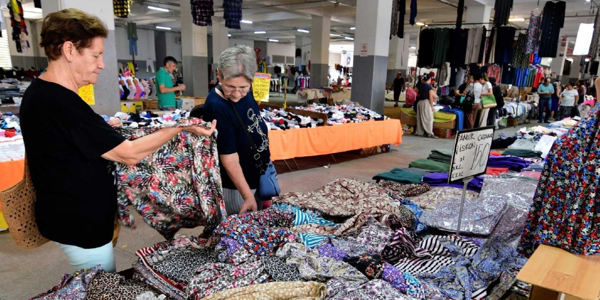 Kapalı bir pazar yerinde, tezgahlardaki kıyafetleri inceleyen iki kadın müşteri. Ön planda, siyah tişörtlü kadınlardan biri elindeki desenli bluzu incelerken, diğeri tezgahtaki ürünlere bakıyor. Tezgahların üzerinde çeşitli renk ve desenlerde katlanmış kıyafetler, askılarda ise elbiseler bulunuyor. Tezgahın üzerindeki tabelada 