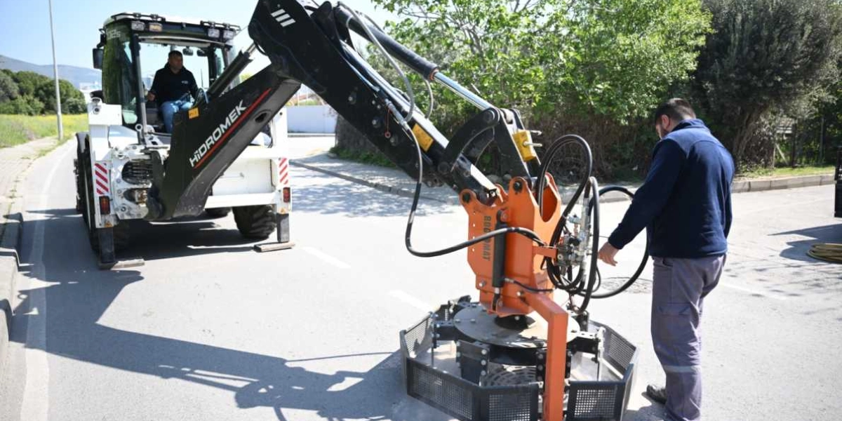 Hidromek marka iş makinesini kullanan bir operatör ve makinenin ucundaki özel aparatla yol çalışması yapan bir işçi, asfalt üzerinde çalışma yürütüyor.