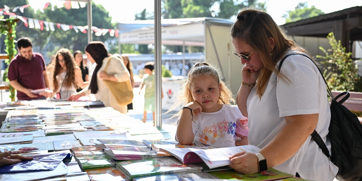 Açık havada kurulan bir kitap fuarı standında, masanın üzerindeki kitapları inceleyen insanlar yer alıyor. Ön planda, beyaz tişörtlü bir kadın ve yanındaki küçük kız çocuğu, masadaki bir kitabı dikkatle inceliyor. Arka planda ise kitaplara bakan diğer ziyaretçiler ve standın üzerinde asılı süslemeler görülüyor.