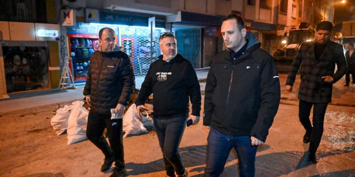 Gece vakti bir sokakta yürüyen dört erkek, devam eden yol çalışmalarını incelemek üzere ilerliyor. Arka planda dükkan vitrinleri ve yol kenarında beyaz çuvallar görülüyor.