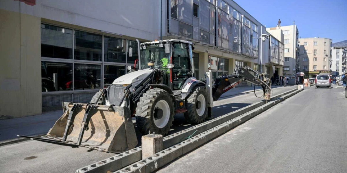Şehir merkezindeki bir caddede, kaldırım kenarında çalışma yapan Hidromek marka beyaz ve siyah renkli bir iş makinesi. Makinenin ön kısmında geniş bir kepçe, arka kısmında ise zemini kırmaya yarayan bir delici uç bulunuyor. Yol kenarına dizilmiş beton bloklar ve arka planda çok katlı binalar görülüyor.