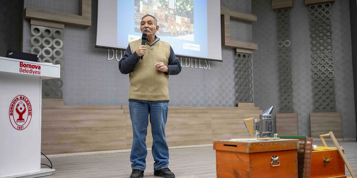 Bornova Belediyesi'ne ait bir konferans salonunda, üzerinde Bornova Belediyesi logosu bulunan kürsünün yanında duran bıyıklı bir adam, elinde mikrofonla konuşma yapıyor. Arka plandaki projeksiyon ekranında arı kovanlarının olduğu bir görsel yer alırken, konuşmacının sağ tarafındaki masanın üzerinde ahşap bir arı kovanı, körük ve arıcılık ekipmanları bulunuyor.