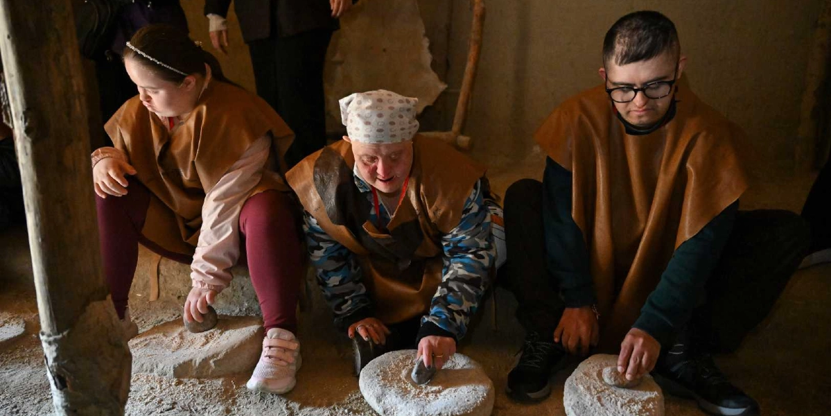 Down sendromlu üç genç, üzerlerinde deri görünümlü kahverengi pelerinlerle, toprak bir zeminde oturmuş geleneksel yöntemlerle taş üzerinde tahıl öğütüyor. Soldaki genç kız saç bandı takmış, ortadaki kişi desenli bir başörtüsü kullanıyor, sağdaki genç ise gözlük takıyor. Hepsi dikkatli bir şekilde ellerindeki taşlarla öğütme işlemini gerçekleştiriyor.