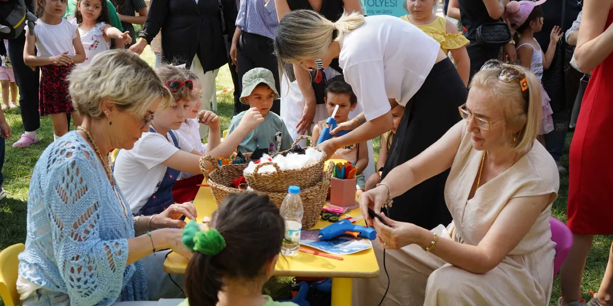 Açık havada, çimenlik bir alanda kurulan sarı bir masanın etrafında toplanmış çocuklar ve onlara yardımcı olan yetişkinler el işi etkinliği yapıyor. Masanın üzerinde hasır sepetler içinde el işi malzemeleri, silikon tabancaları ve renkli çubuklar bulunuyor. Yetişkinler, çocukların yaratıcı çalışmalarına rehberlik ederken, çevrede etkinliği izleyen diğer çocuklar ve aileler yer alıyor.