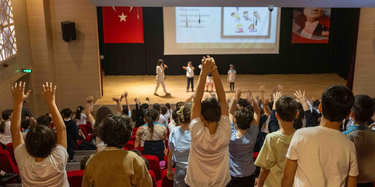 Konferans salonunda sahnedeki bir eğitmen ve çocukların eşliğinde, koltuklarda oturan çok sayıda öğrencinin ellerini havaya kaldırarak katılım gösterdiği bir sağlık eğitimi etkinliği. Sahnedeki dev ekranda egzersiz hareketlerini gösteren çizimler yer alırken, arka planda Türk bayrağı ve Atatürk posteri asılı duruyor.
