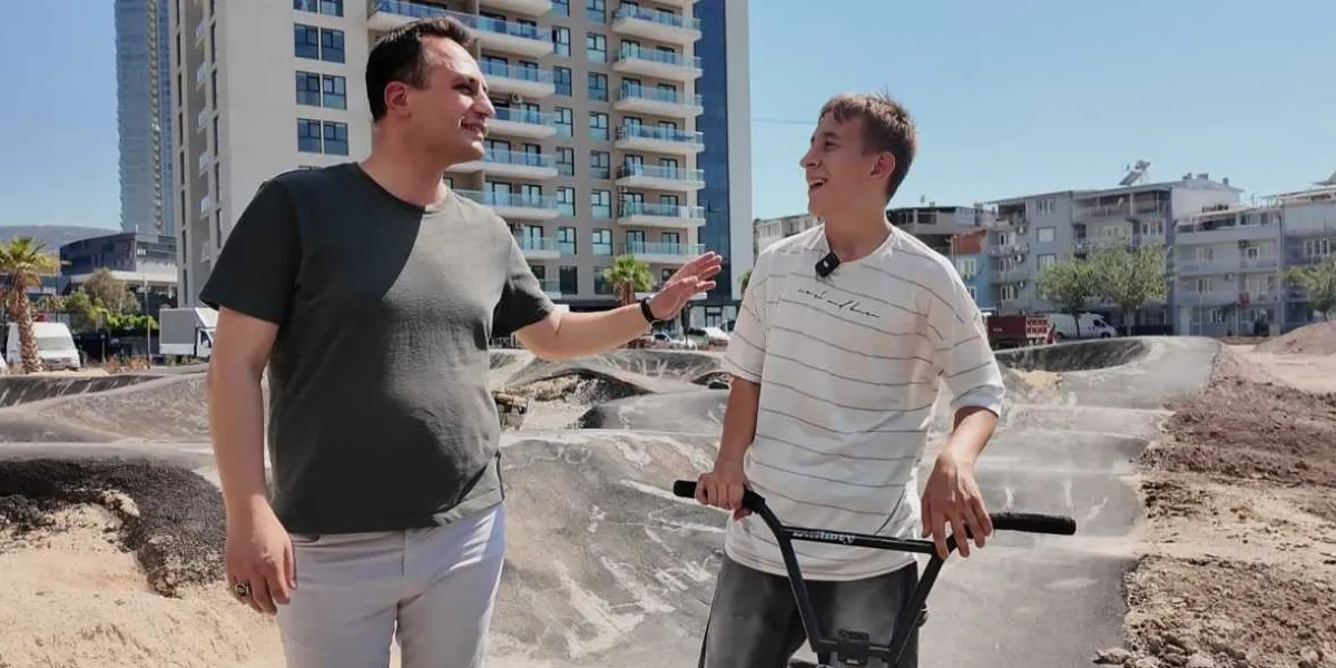 Bornova'da yapımı devam eden bir bisiklet parkurunda, gri tişörtlü bir yetişkin ile elinde BMX bisikleti tutan genç bir bisikletçi sohbet ediyor. Arka planda modern apartmanlar ve toprak zeminli, engebeli bir sürüş alanı görülüyor.