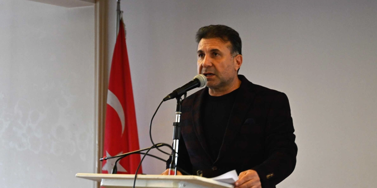 Koyu renkli ceket ve siyah tişört giymiş orta yaşlı bir erkek, kürsüde mikrofon başında konuşma yapıyor. Arka planda bir Türk bayrağı yer alıyor.