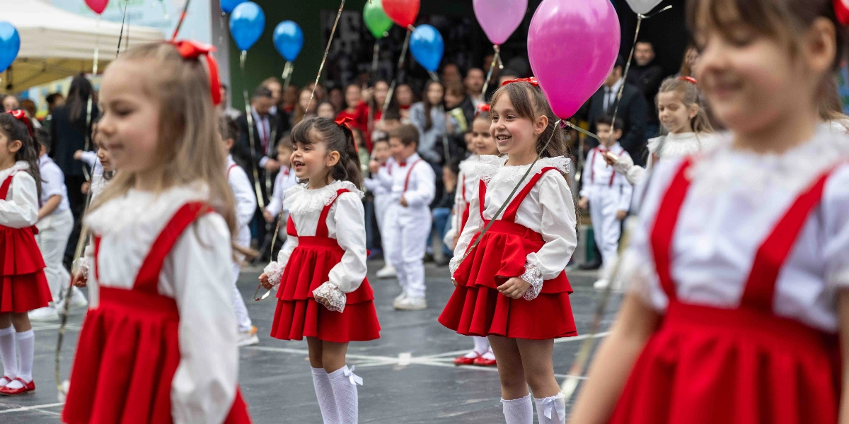 Beyaz gömlek ve kırmızı askılı etek giymiş kız çocukları ile beyaz kıyafetli erkek çocuklarının ellerinde renkli balonlarla katıldığı bir kutlama töreni. Çocuklar neşeyle yürürken arka planda yetişkinler izleyici olarak yer alıyor.