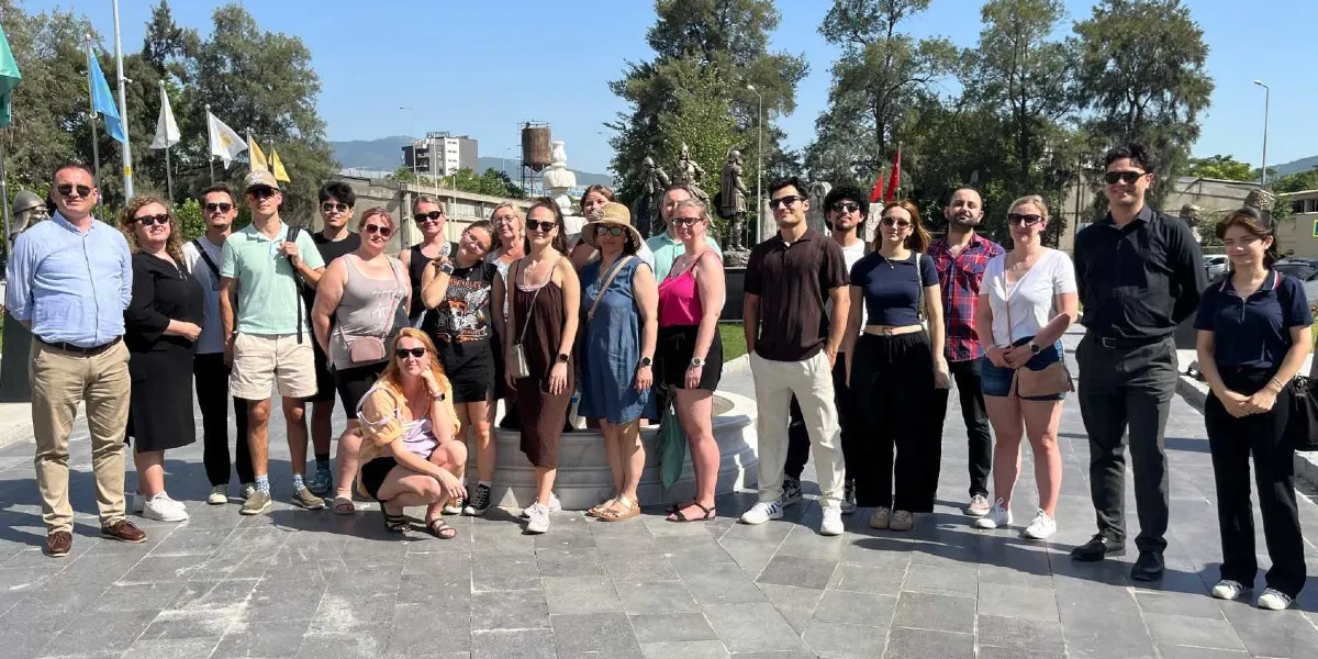 Açık havada, güneşli bir günde park benzeri bir alanda bir araya gelmiş, farklı yaş ve giyim tarzlarına sahip yaklaşık yirmi kişilik genç ve yetişkin grubu. Gruptakilerin çoğu güneş gözlüğü takıyor ve kameraya gülümseyerek poz veriyor. Arka planda ağaçlar, bayrak direkleri ve bir heykel figürü seçiliyor. Grubun bir kısmı ayakta, bir kısmı ise ön tarafta bir çeşme yapısının etrafında duruyor veya çömelmiş durumda.