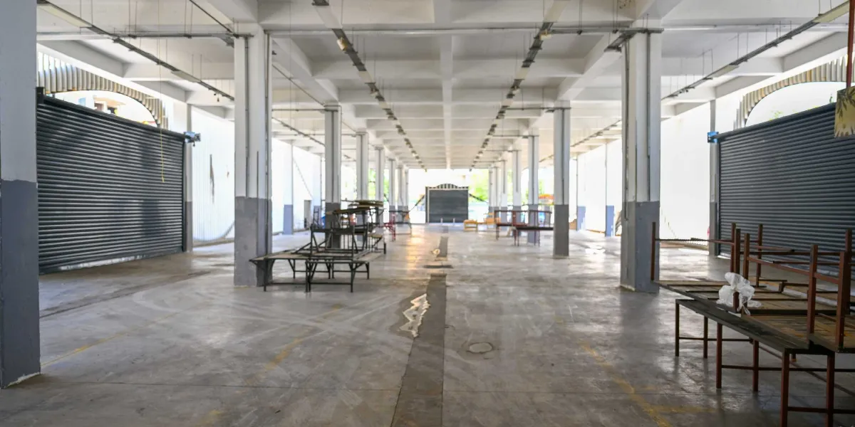 Kapalı, geniş ve boş bir pazaryeri alanı. Beton zemin üzerinde, tezgahların kurulduğu metal iskeletler dağınık bir şekilde duruyor. Yan duvarlarda kapalı metal kepenkler bulunuyor ve tavan yüksek sütunlarla destekleniyor.