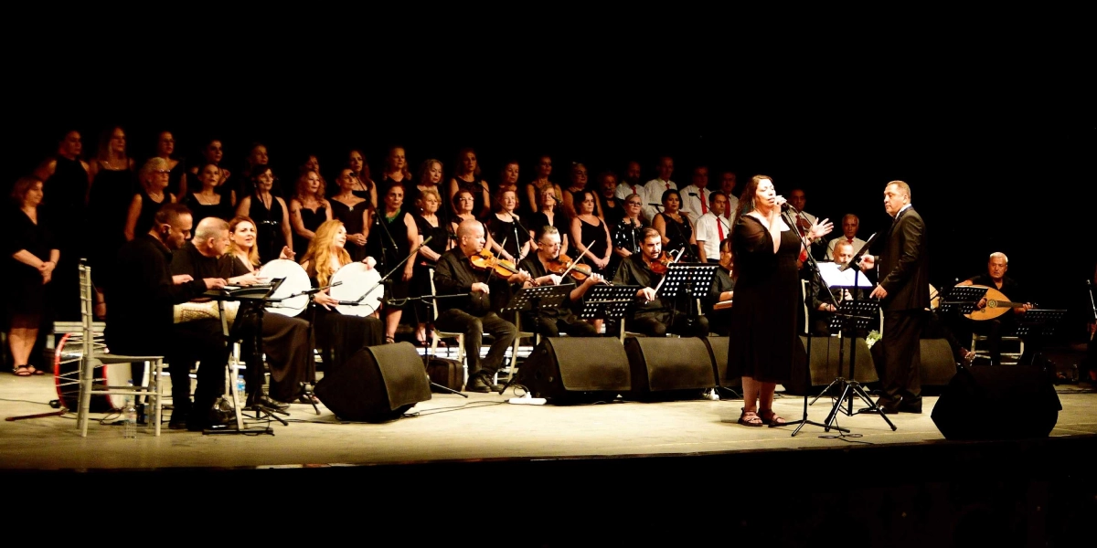 Siyah bir sahnede, siyah kıyafetler giymiş kalabalık bir koro, yaylı çalgılar grubu ve solistlerin yer aldığı bir konser performansı. Sahnenin ön kısmında ayakta şarkı söyleyen kadın solist ve yanında duran erkek şef bulunurken, arkalarında keman çalan müzisyenler, ellerinde tef tutan kadınlar ve arka planda geniş bir koro grubu yer alıyor. Sahne üzerinde müzik sehpaları, mikrofonlar ve ses sistemleri bulunuyor.