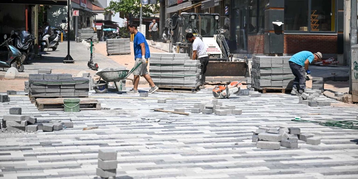 Bornova Belediyesi ekipleri, bir cadde üzerinde gri ve beyaz taşlarla yeni kaldırım döşeme çalışması yapıyor. İşçilerden biri el arabasıyla malzeme taşırken, diğerleri istiflenmiş taşları yerleştiriyor ve bir iş makinesi çalışma alanında bekliyor.